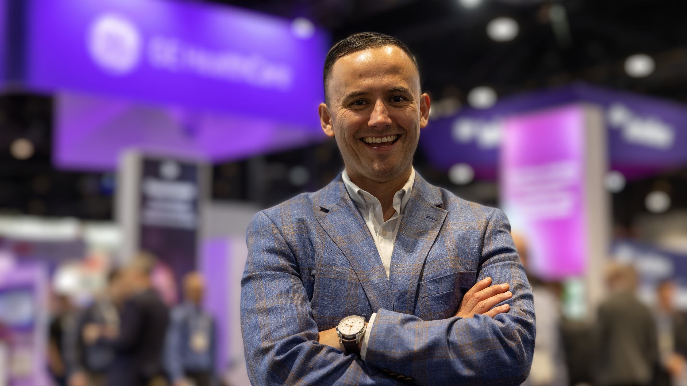 Saul Marquez stands confidently with arms crossed at a healthcare conference, wearing a blue plaid blazer and white shirt. The blurred background features purple signage and booths, suggesting a vibrant industry event focused on digital health or med tech.