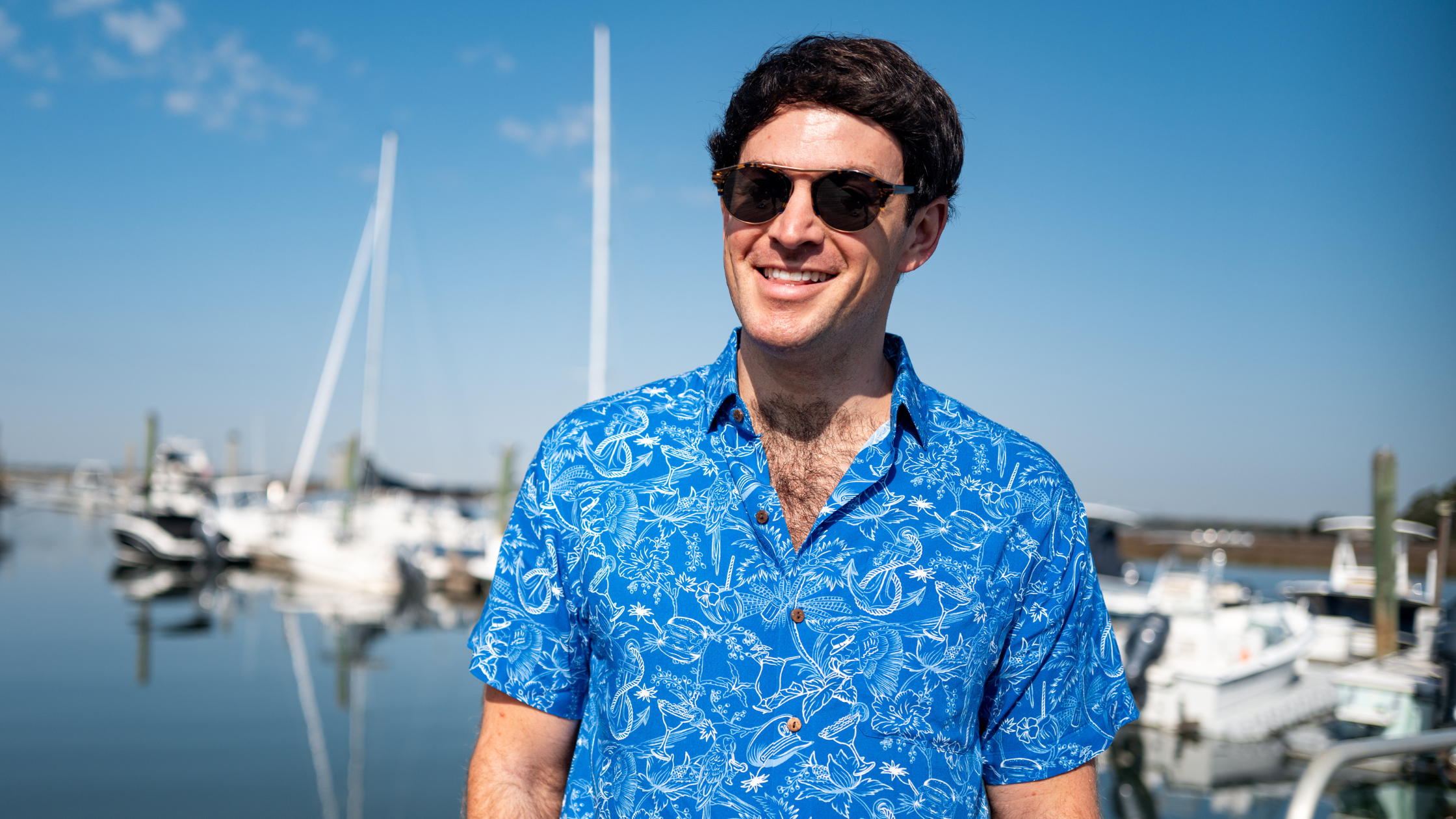 Kenny Haisfield, founder of Kenny Flowers, smiles while standing at a marina on a sunny day. He’s wearing a vibrant blue short-sleeve shirt with white tropical prints and dark sunglasses. Sailboats and clear blue skies fill the background, giving the image a relaxed, coastal vibe.