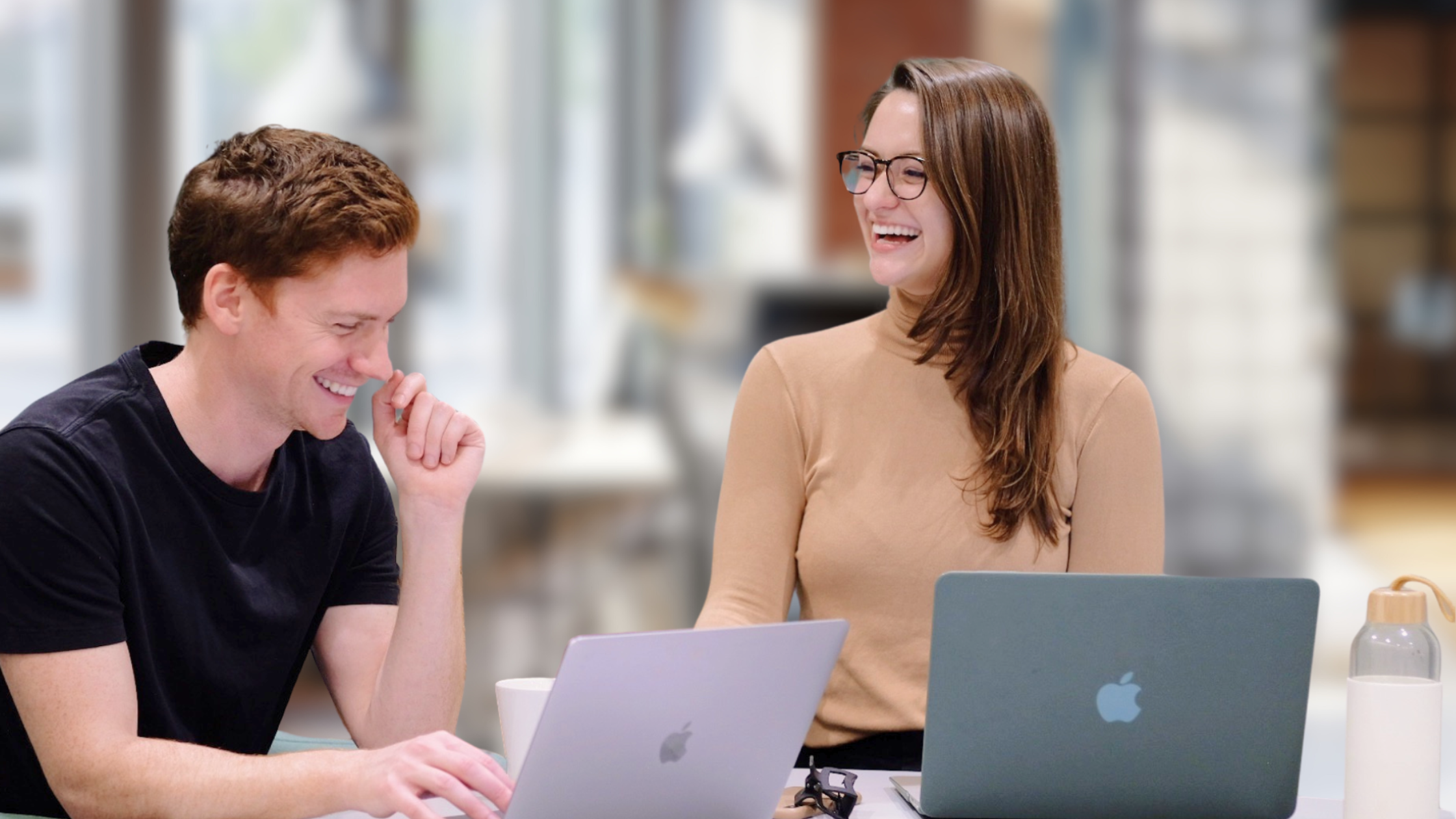 Deirdre and Dave Purdy laugh together while working at a desk with their laptops. Dave, in a black t-shirt, looks down while smiling, and Deirdre, wearing glasses and a tan sweater, looks toward him mid-laugh. A blurred modern office background suggests a collaborative, energetic work environment.